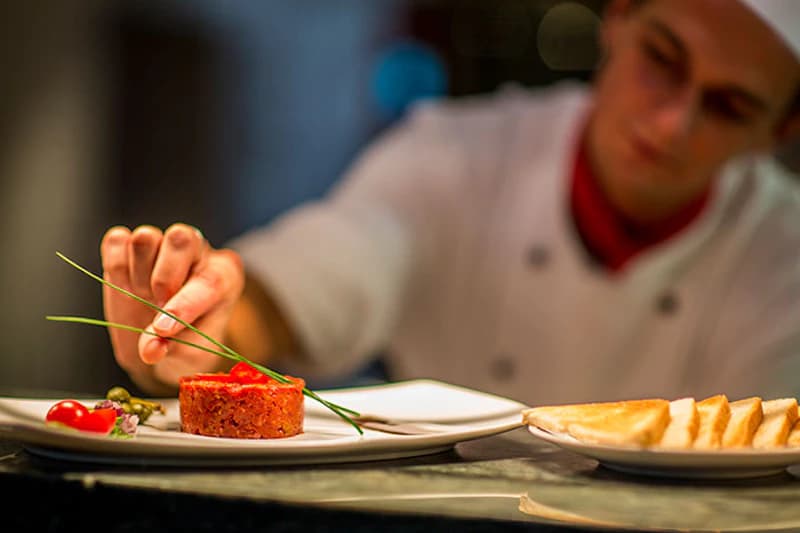 Koch in weißer Jacke legt Schnittlauch auf Tartar; Kirschtomaten, Kapern und ein Teller mit Toastscheiben sichtbar.