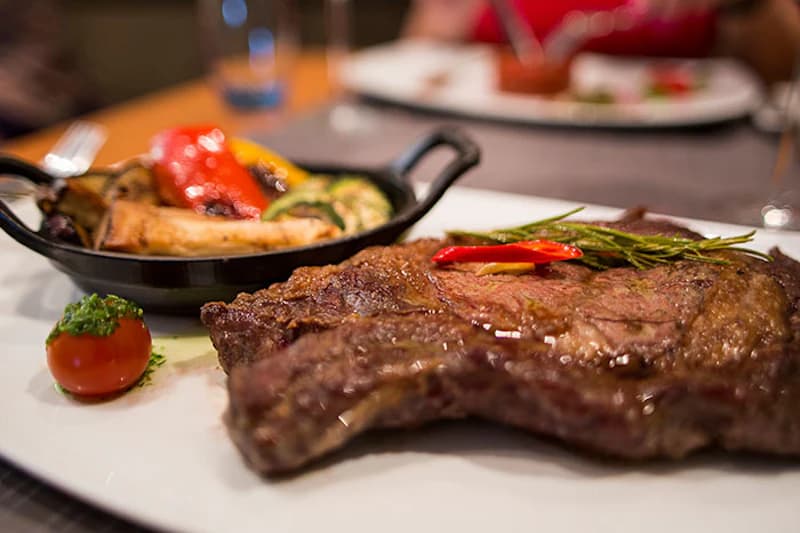 Gegrilltes Steak auf Teller mit Rosmarin, Chiliring, Pfännchen mit gebratenem Gemüse und Kräutertomate.