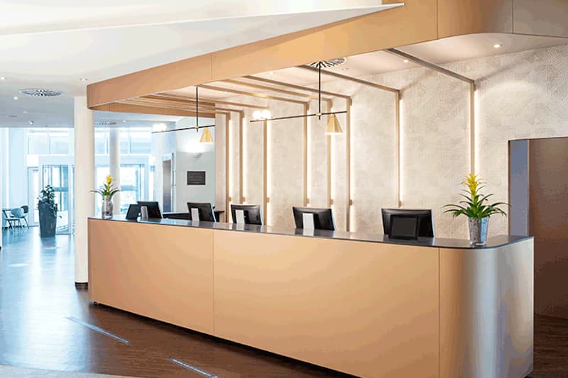 Helle Hotelrezeption mit langem Tresen, vier Computermonitoren, Hängeleuchten und Pflanzen, Eingangsbereich hinten.