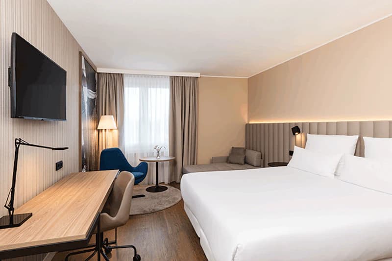 Hotelzimmer mit Doppelbett, Schreibtisch, Wandfernseher und Sitzbereich mit blauem Sessel am Fenster.