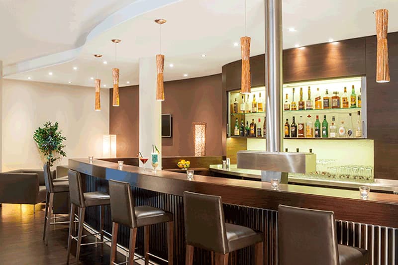 Hotelbar mit langer Holztheke, Barhockern, beleuchteten Spirituosenregalen und Hängeleuchten.