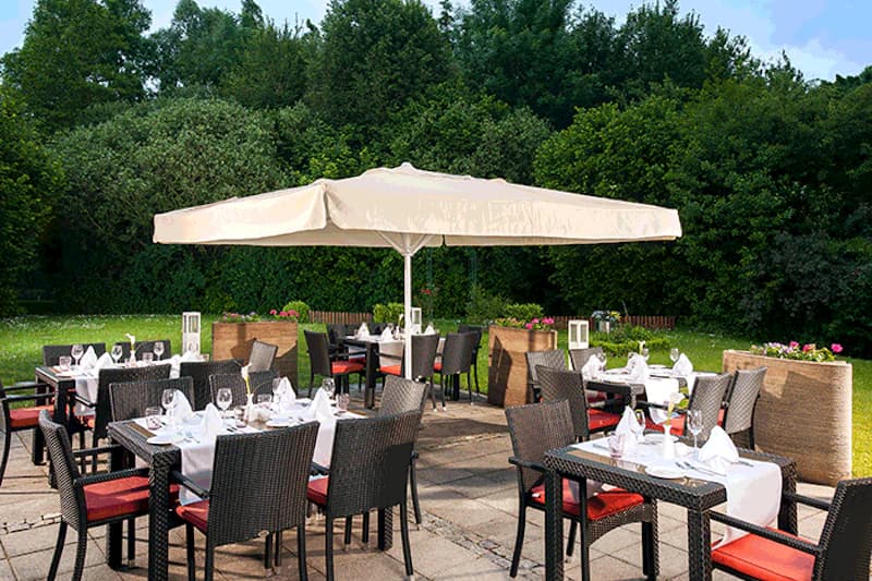 Außen-Terrasse eines Hotelrestaurants mit gedeckten Tischen, Sonnenschirm und Stühlen vor dichtem Baumbestand.