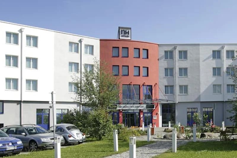 Dreigeschossiges Hotelgebäude mit rotem Mittelteil, NH-Logo auf dem Dach, Eingangsbereich, parkende Autos und Rasen.