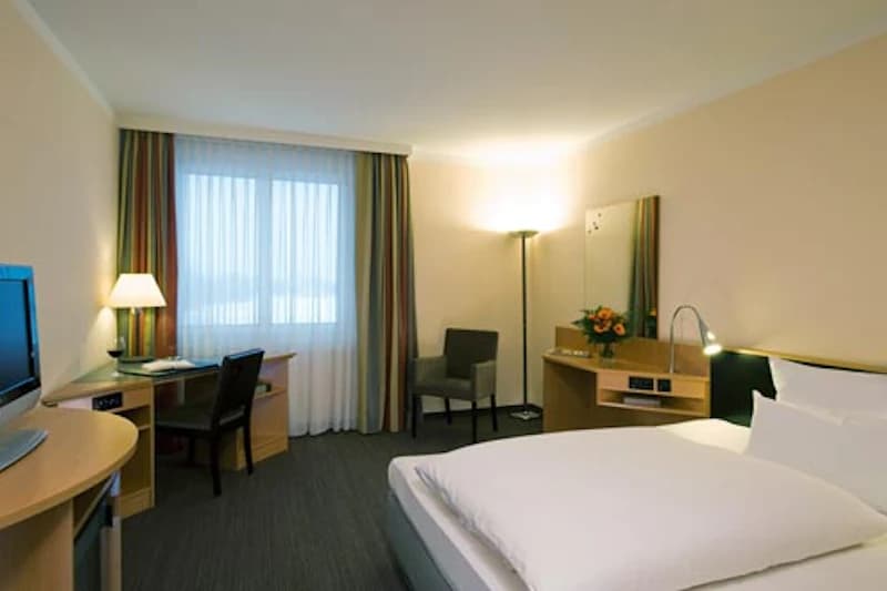 Hotelzimmer mit Doppelbett, Nachttischlampe, Schreibtisch, Fernseher, Sessel und Vorhängen.
