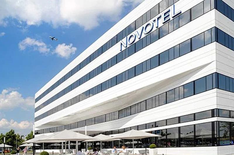 Weiße Hotelfassade mit Novotel-Schriftzug, langen Fensterreihen, Sonnenschirmen auf der Terrasse und Flugzeug am Himmel.