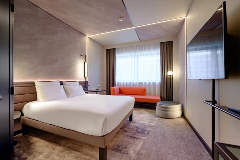 Modernes Hotelzimmer mit Doppelbett, orangefarbener Couch am Fenster, Wandfernseher und indirekter Beleuchtung.