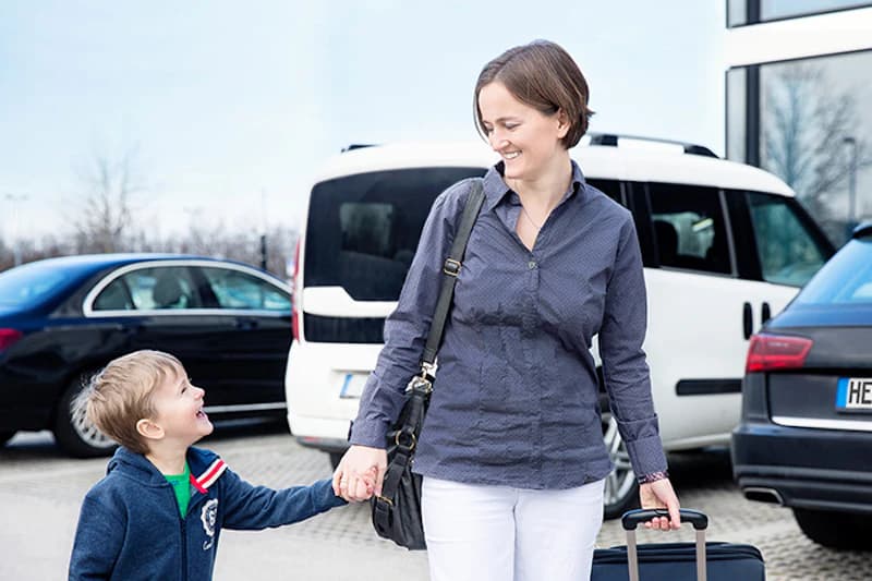 Frau zieht Trolley und hält die Hand eines kleinen Jungen auf einem Parkplatz vor parkenden Autos.