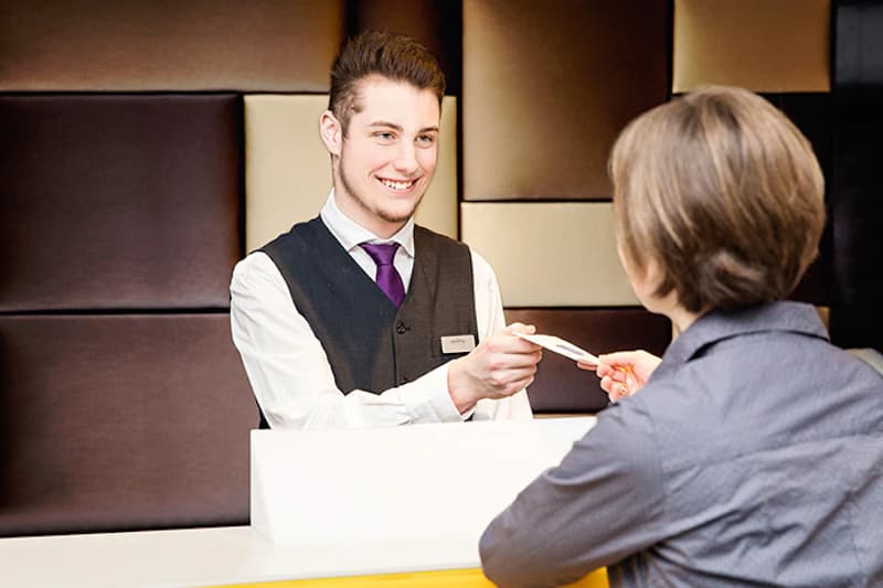 Junger Hotelmitarbeiter reicht einer Gästin an der Rezeption eine Schlüsselkarte.