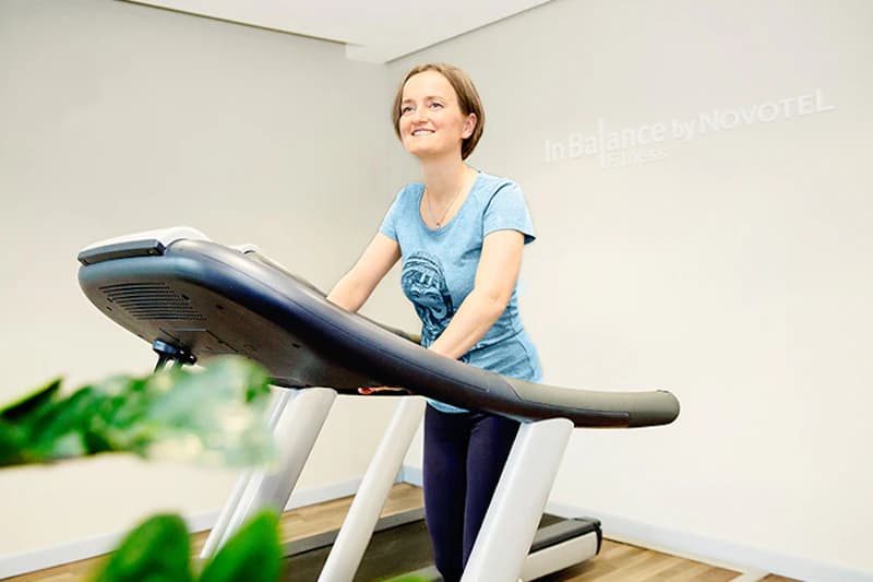 Frau auf Laufband im Hotelfitnessraum, Wandschrift InBalance by NOVOTEL im Hintergrund.