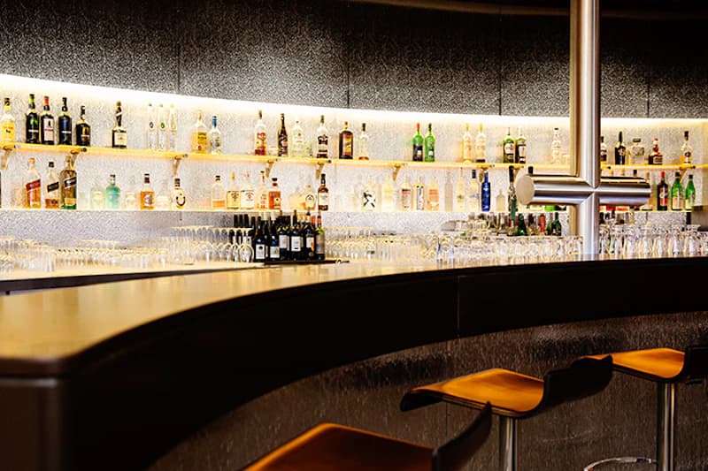 Hotelbar mit beleuchteten Regalen voller Spirituosen, vielen Gläsern und Tresen mit Barhockern.