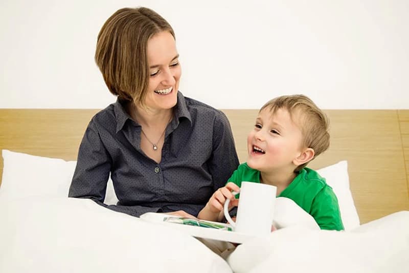 Mutter und kleiner Sohn sitzen im Bett und lachen, Sohn hält eine Tasse auf einem Tablett.