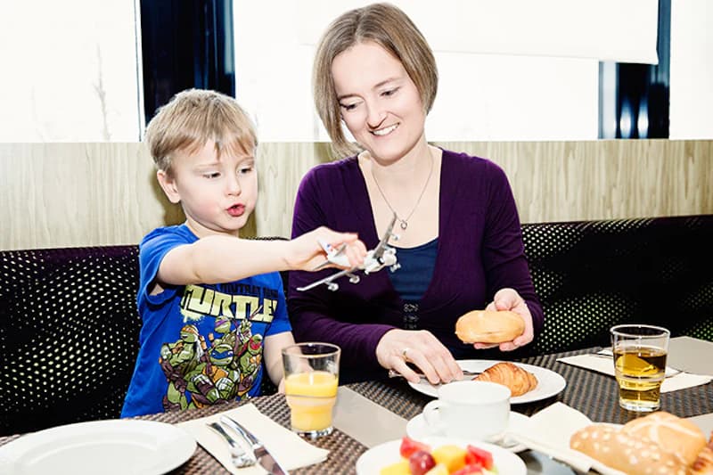 Mutter und Sohn am Tisch, Kind spielt mit Spielzeugflugzeug, auf dem Tisch Gebäck, Orangensaft und Tasse.