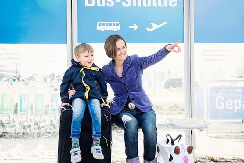 Frau und Kind sitzen auf Koffer an Haltestelle mit blauem Schild und Bus- und Flugzeugpiktogramm.