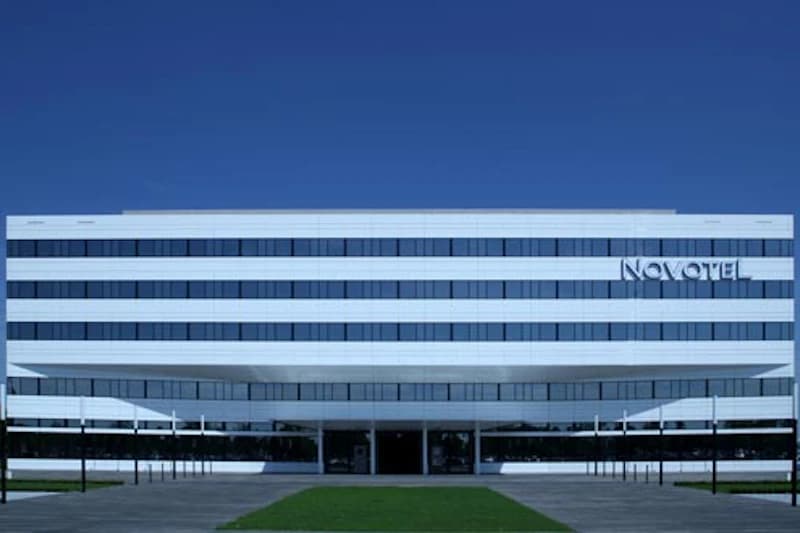 Fassade des Novotel München Airport mit weißen horizontalen Bändern, Reihenfenstern und Eingangsüberdachung vor Rasen.