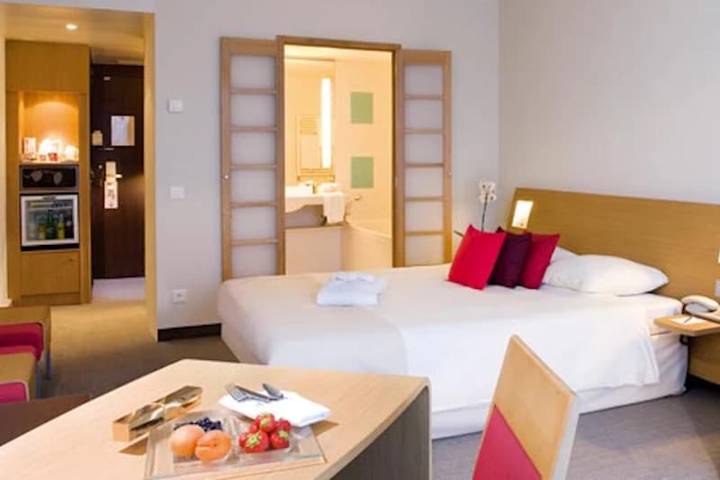 Hotelzimmer mit Doppelbett, roten Kissen, Tisch mit Obstplatte, geöffneter Schiebetür zum Bad und Minibar beim Eingang.