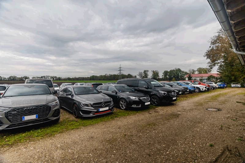 Reihe geparkter Autos auf unbefestigtem Platz neben Feld und Strommast unter bewölktem Himmel.