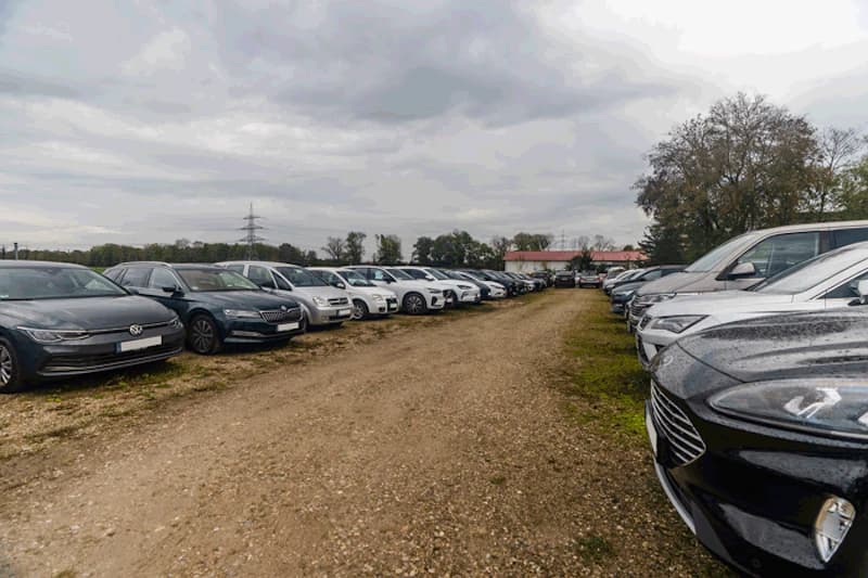 Zwei Reihen geparkter Autos auf geschottertem Feldweg bei bewölktem Himmel, Bäume und Gebäude im Hintergrund.