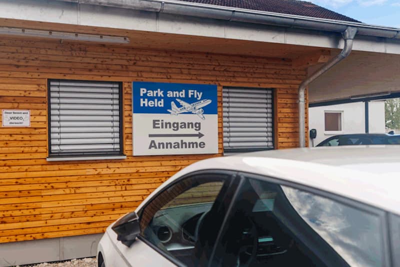 Holzfassade eines Parkplatzbüros mit Schild Park and Fly Held Eingang Annahme und weißem Auto im Vordergrund.