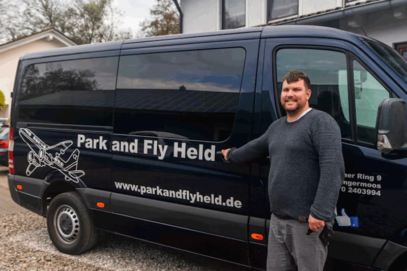 Mann steht neben dunkelblauem Bus mit Schriftzug Park and Fly Held und zeigt auf die Tür.