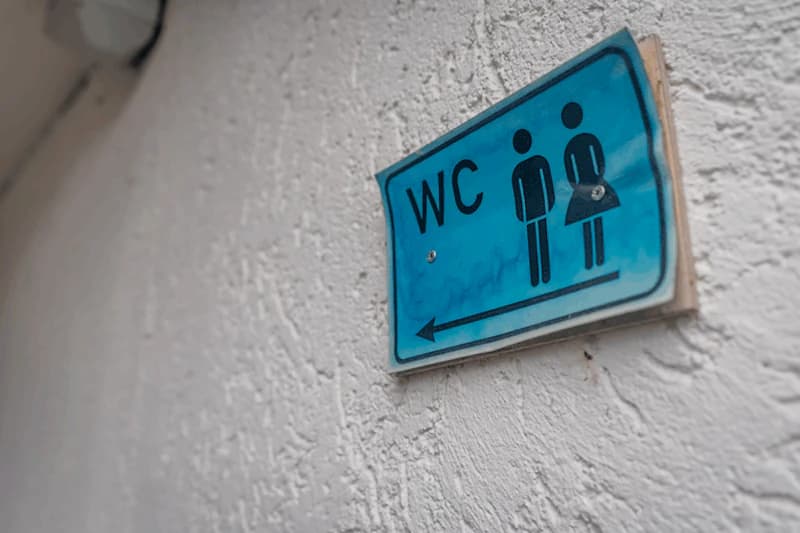 Blaues WC-Schild mit Männer- und Frauensymbol und Pfeil nach links an weißer Wand.