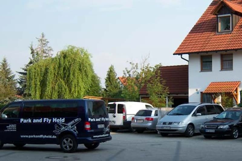 Parkplatz mit mehreren Autos und blauem Shuttlebus Park and Fly Held vor einem Wohnhaus.