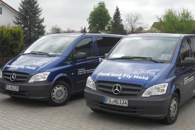 Zwei blaue Mercedes Kleinbusse mit Aufschrift Park and Fly Held auf gepflastertem Stellplatz vor Wohnhäusern.
