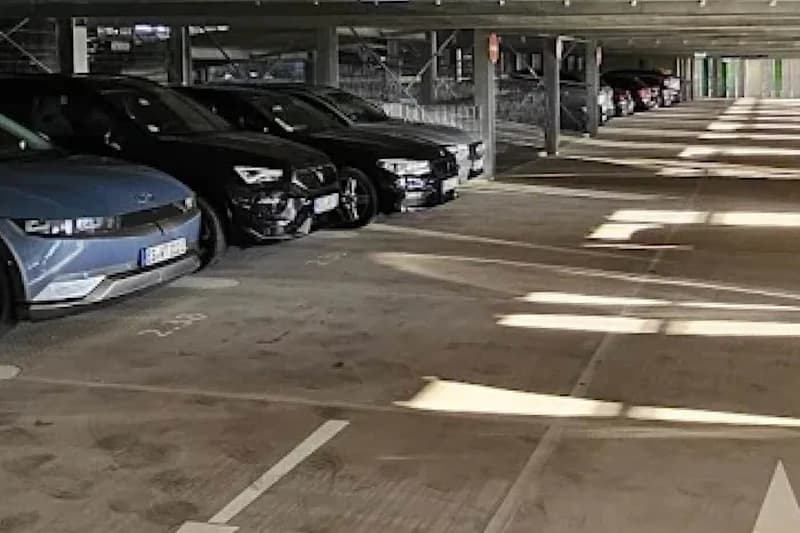 Überdachtes Parkhaus mit geparkten Autos links, Betonstützen und Lichtflecken auf dem Boden.