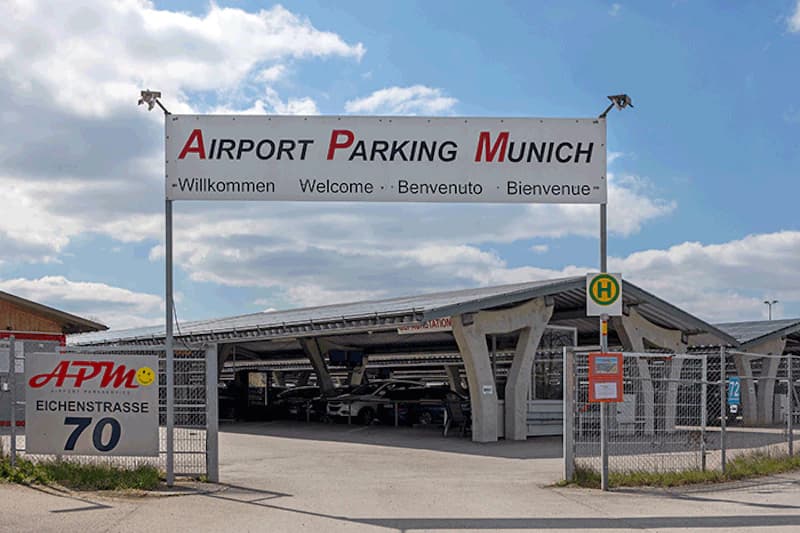 Eingang zu einem umzäunten Parkplatz mit großem Schild Airport Parking Munich und überdachten Parkreihen.