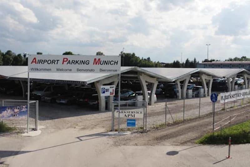 Umzäunter Carport-Parkplatz mit überdachten Stellreihen, Einfahrt mit Schild Airport Parking Munich und geparkten Autos.