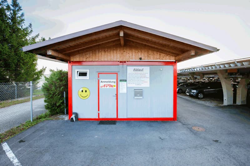Kleiner Anmeldekiosk mit Holzgiebel, grauen Wänden, rotem Rahmen und Smiley-Aufkleber, Zaun links, Carports mit geparkten Autos rechts.