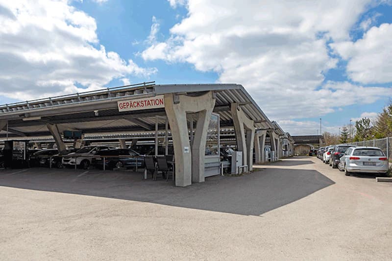 Überdachte Carports mit geparkten Autos und Schild GEPÄCKSTATION, rechts Fahrweg mit weiteren geparkten Wagen und Zaun.