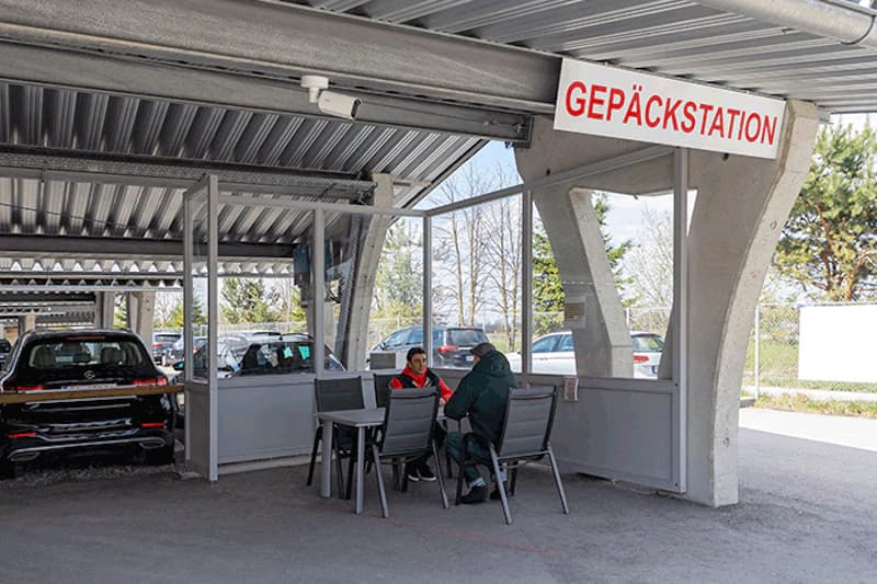 Überdachter Carport mit Gepäckstation, zwei Personen sitzen an Tisch, parkende Autos im Hintergrund.
