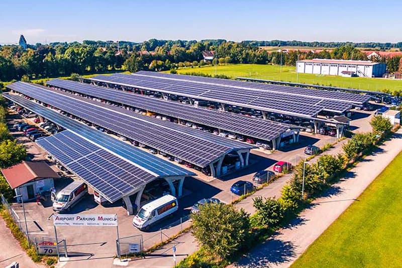 Reihen von überdachten Parkplätzen mit Solarpaneelen auf den Dächern, geparkte Autos und Shuttlebus, umzäuntes Gelände.
