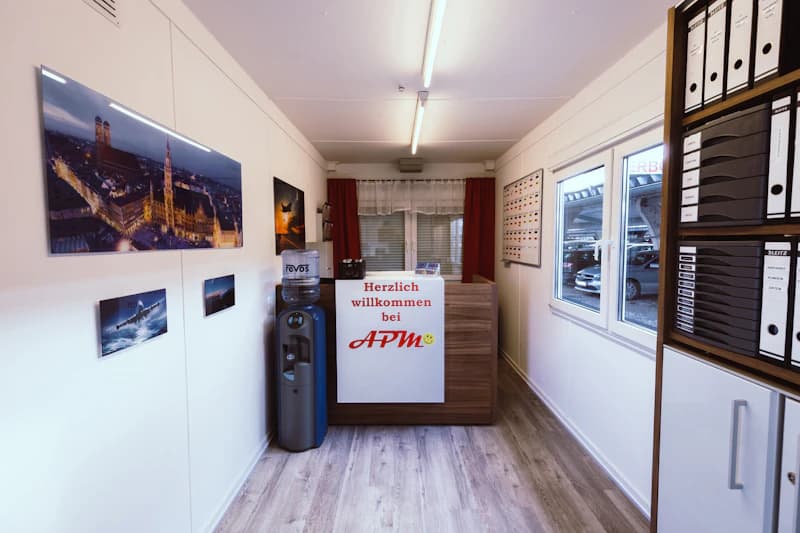 kleiner Empfangsraum mit Tresen Herzlich willkommen bei APM, Wasserspender, Wandbildern, Regal mit Ordnern und Fenster zum Parkplatz.