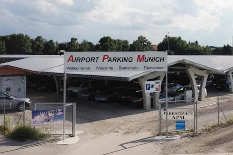 Überdachte Parkplätze unter Carports, großes Schild Airport Parking Munich, mehrere Autos hinter Zaun auf Schotter.
