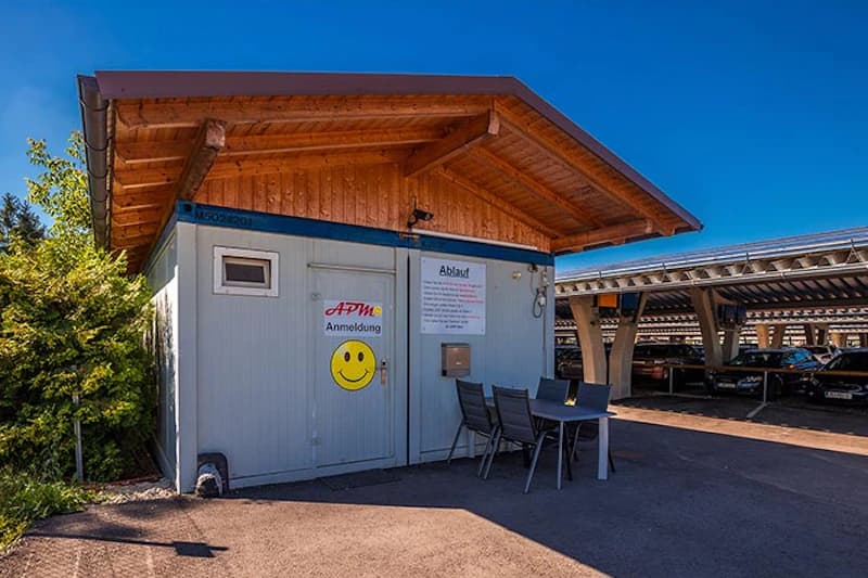 Kleines Anmeldehäuschen mit überdachtem Holzdach, APM Schriftzug und gelbem Smiley; Tisch und Stühle; überdachte Parkplätze im Hintergrund.