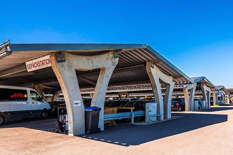 Überdachte Parkreihen mit markanten Betonsäulen, mehreren Autos, APM-Lieferwagen und Mülltonnen neben einer Gepäckstation.