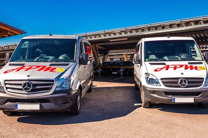 Zwei weiße Mercedes Transporter mit APM Logo vor überdachten Carports auf dem Gelände am Flughafen München.