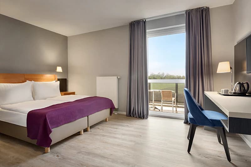 Helles Hotelzimmer mit Doppelbett, violettem Überwurf, Schreibtisch und Balkon mit Stühlen.