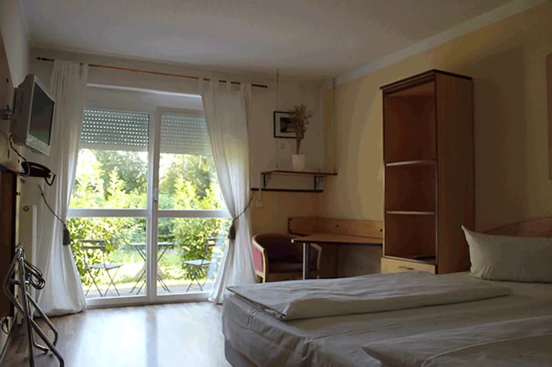 Hotelzimmer mit zwei Einzelbetten, großer Glasfront zur Terrasse mit zwei Stühlen, Schreibtisch, Regal und Wandfernseher.