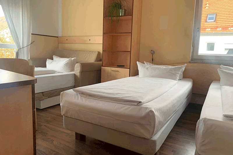 Drei Einzelbetten und ein Sofabett in einfachem, hellem Zimmer mit Regal und Fenster.