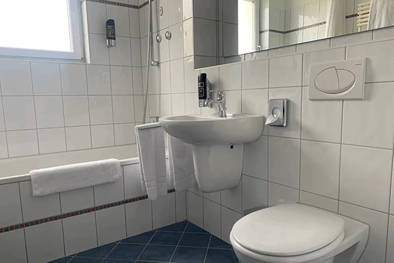 Weiß gefliestes Badezimmer mit Toilette, Wandwaschbecken, Badewanne, Handtüchern und Spiegelschrank.
