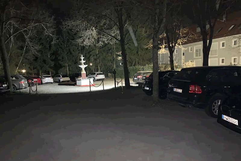 Beleuchteter Parkplatz bei Nacht mit geparkten Autos, einem hell beleuchteten Brunnen und Bäumen neben einem Wohngebäude.