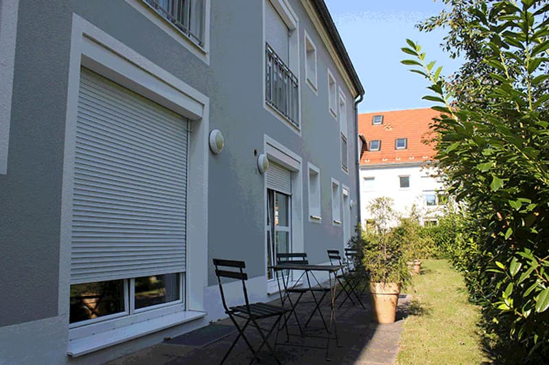 Außenfassade eines hellgrauen Gebäudes mit geschlossenen Rollläden, Terrasse mit Klappstühlen und Pflanzkübeln, Hecke.