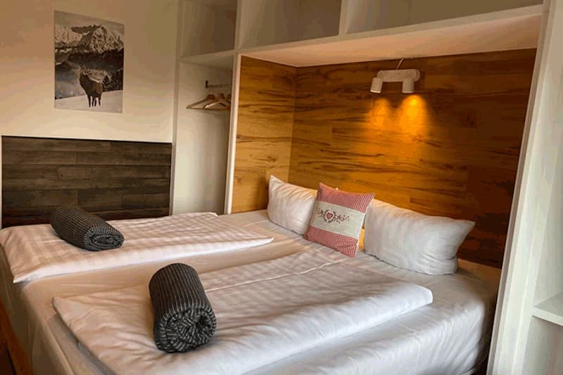 Zwei Betten mit weißer Bettwäsche, gerollten Handtüchern, Kissen, warm beleuchteter Holzrückwand, offenem Kleiderschrank und Wandbild.