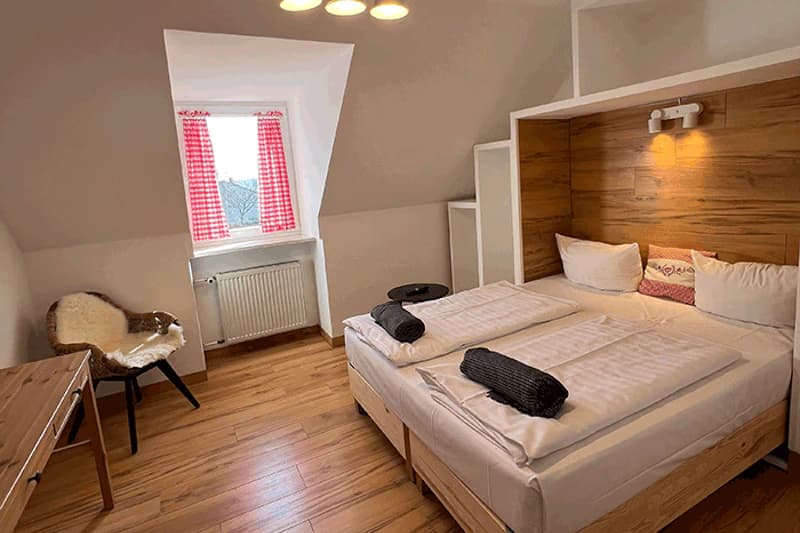 Dachzimmer mit zwei Einzelbetten, Holzkopfteil, Nachtlicht, Giebelfenster mit rotkarierten Vorhängen und Stuhl mit Fell.
