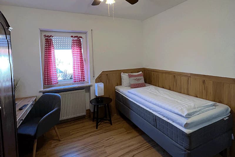 Einzelzimmer mit Einzelbett, Holzwandverkleidung, kleinem Nachttisch mit Lampe, Schreibtisch und karierten Vorhängen.