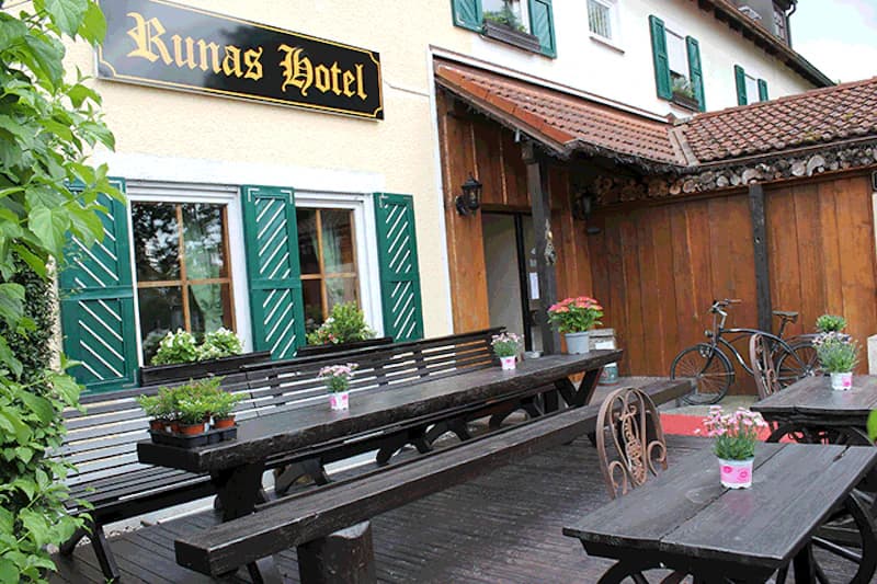 Außenbereich mit Holztischen und Bänken, Blumentöpfen, Fahrrad vor Fassade mit Runas Hotel Schild.