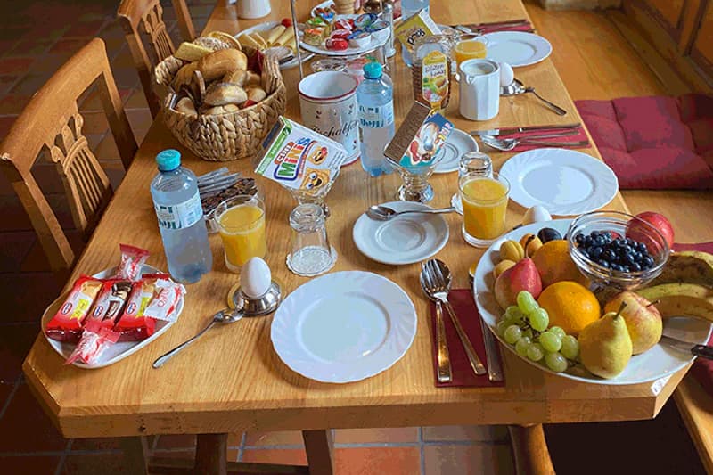 gedeckter Frühstückstisch mit Tellern, Brötchenkorb, Obstschale, Orangensaft, Wasserflaschen und Eiern.