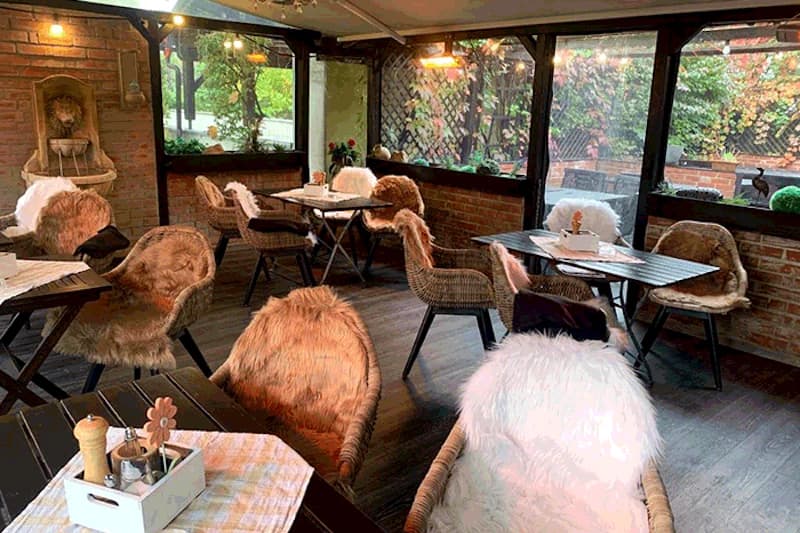 Überdachte Hotelterrasse mit Korbsesseln, Fellüberwürfen, Holztischen und Ziegelwand, Fenster zum begrünten Außenbereich.
